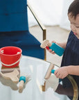 Child playing with wooden Cleaning Set, featuring a bucket, broom, spray-bottle, and dustpan on a glass table.