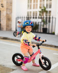 Child riding the Micro Balance Bike Flamingo Pink on a sunny day, showcasing fun and safety with a helmet and knee pads.