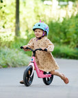Child riding a Micro Balance Bike Flamingo Pink, wearing a helmet, enjoying a sunny day in the park.