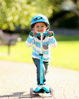 Child riding Mini Micro Deluxe scooter outdoors, wearing a helmet and smiling in bright sunlight.