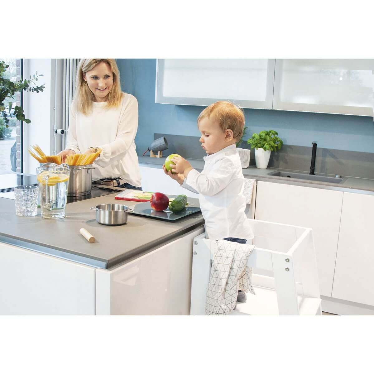 A child standing in the kitchen on a learning tower cutting fruit with mother