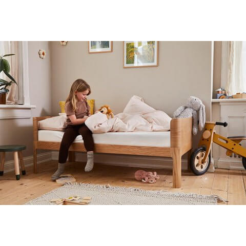 Child sitting on FLEXA Nova Junior Bed in a cozy room with toys and soft bedding, featuring an oak finish.