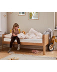 Child sitting on FLEXA Nova Junior Bed in a cozy room with toys and soft bedding, featuring an oak finish.