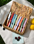 KiddiKutter Knife - Cotton Candy collection of colorful knives in a woven basket with fresh fruits on a picnic blanket.