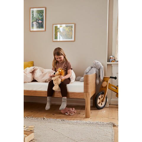 A young girl sitting on a FLEXA Nova Junior Bed, playing with toys in a cozy, well-decorated kids&#39; room.