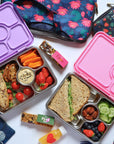 Yumbox Pret stainless steel lunch box with colorful compartments, vegetables, fruits, and snacks inside a vibrant lunch setting.